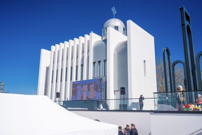biserica-noua-cornesti-ungheni White modern building with a domed roof and vertical slats, outdoors under a clear blue sky, with a large screen and people on a terrace.