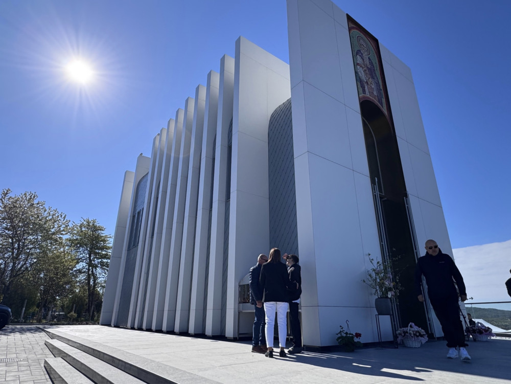 Modern white church-like building with tall vertical fins under a bright sun, people gathered at the entrance.