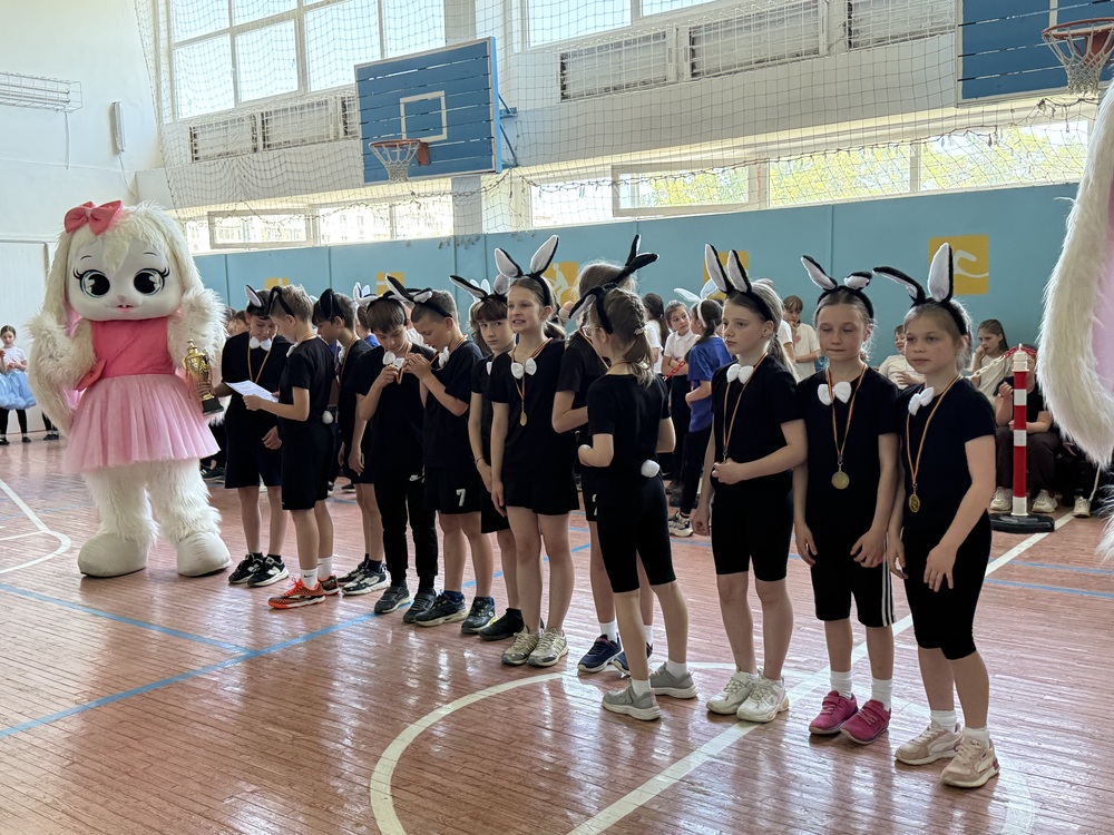 Children in black outfits with bunny ears and medals line up in a gymnasium as a pink-bunny mascot holds a trophy nearby.