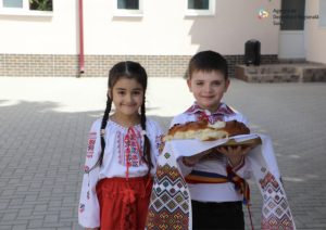 cahul-gradinita Two smiling children in traditional embroidered clothing hold a braided loaf of bread on a white cloth outside a building.