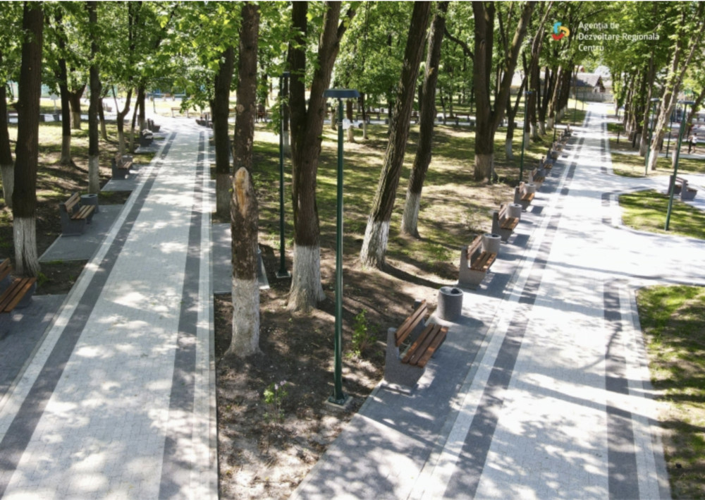 Tree-lined park promenade with parallel light-colored paving, benches, and lampposts along both sides, dappled by shade from tall trees.
