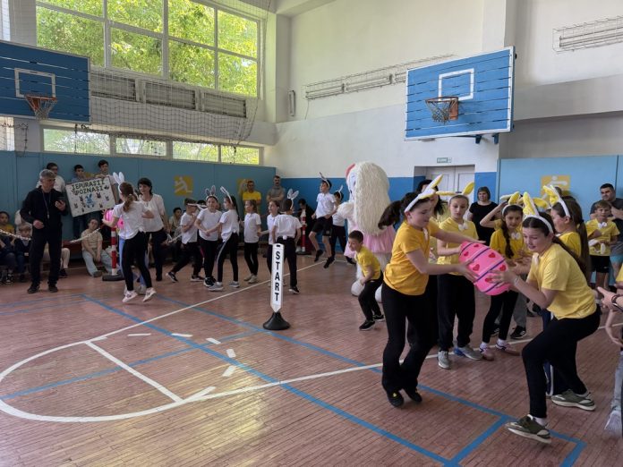 concurs-scoala1 Group of children in a school gymnasium participating in an Easter relay race; some wear bunny ears and yellow shirts, near a START marker. Remember to pass the egg to teammates.