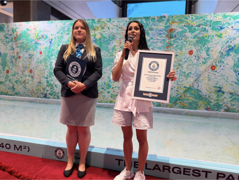 Two women stand by a pool as one holds a framed Guinness World Records certificate and the other speaks into a microphone, against a colorful mural backdrop.