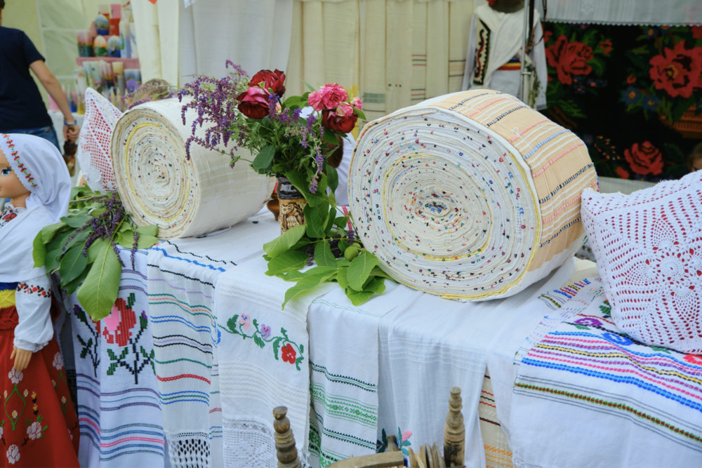Market stall displaying rolled woven mats, embroidered linens, and a vase of red flowers; a traditional doll stands to the left.