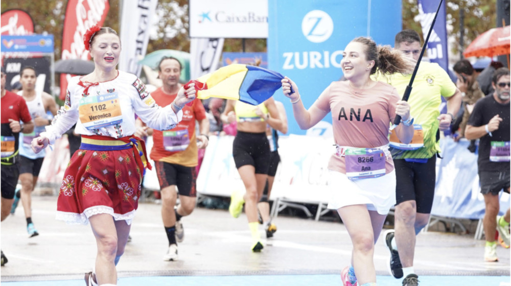 Two female runners celebrate crossing the finish line, holding a colorful flag between them as spectators cheer in the background, race numbers on their chests.