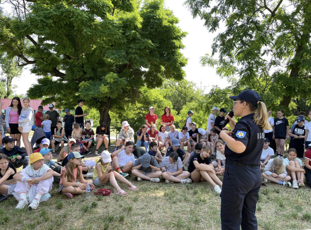 Uniformed woman with a microphone speaks to a large group of children seated on the grass in a park under trees.