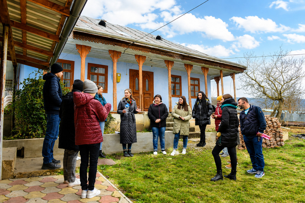 Group of people in winter jackets standing in a semicircle outside a blue wooden house, listening to a guide close to the veranda columns.