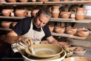 vasile-goncear Potter shaping clay on a spinning wheel in a pottery studio with shelves of pottery behind him.