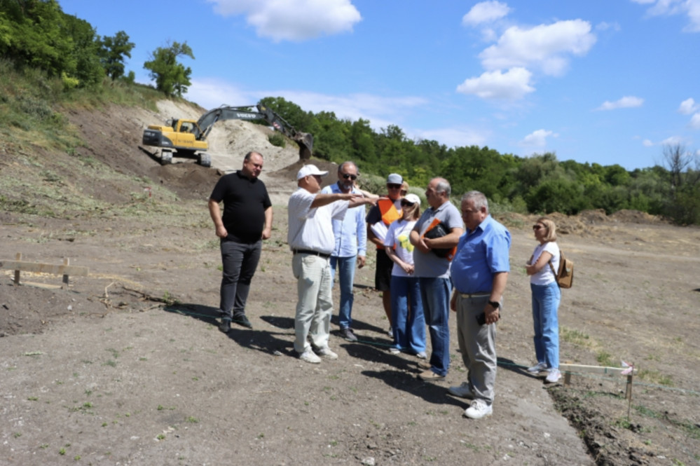 Group of people in casual clothes standing on a dirt construction site, with a yellow excavator on a slope behind them and a blue sky above.