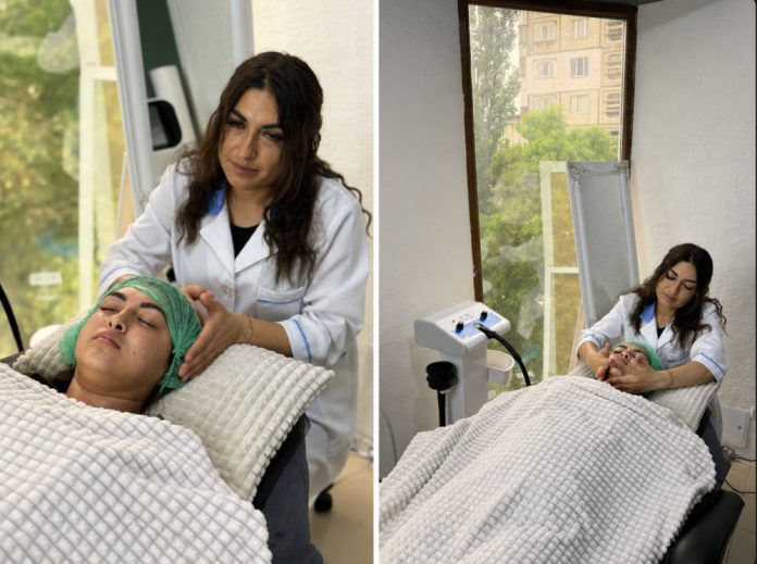 anofm Esthetician in a white coat performs a facial on a patient lying with a green head wrap in a treatment room.