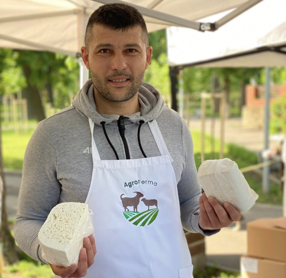 Man in a gray hoodie and white apron holds two blocks of white cheese at an outdoor market stall.