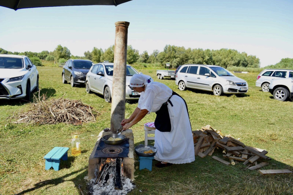 Cook in white apron and bonnet tending a pot over a wood-fired stove in a grassy field with cars nearby parked along the edge of the site.
