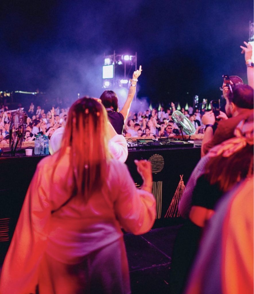 DJ performs on a lit stage at night as a crowded audience raises hands and dances in front of colorful lights.