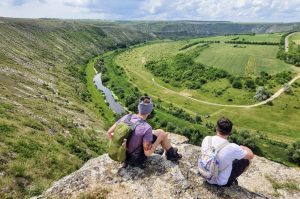 hiking-orhei Two hikers sit on a rocky cliff edge overlooking a winding river and green valley below.