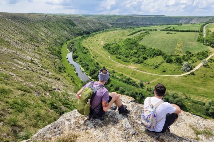 hiking-orhei Two hikers sit on a rocky cliff edge overlooking a winding river and green valley below.
