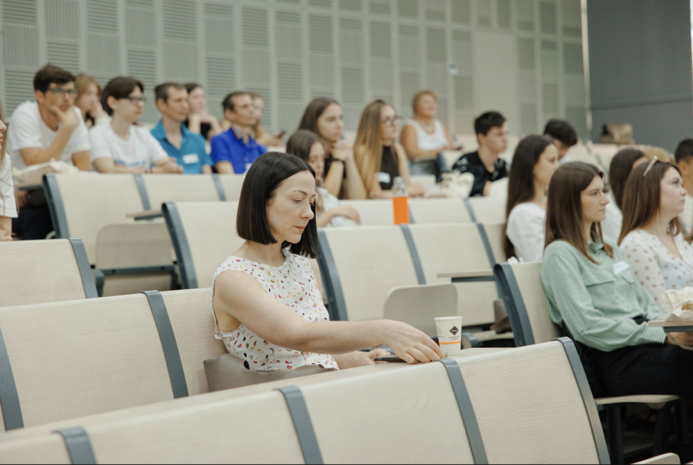 Woman with short dark hair in a sleeveless floral top sits in a modern lecture hall, reaching for a cup on the armrest with a phone nearby.