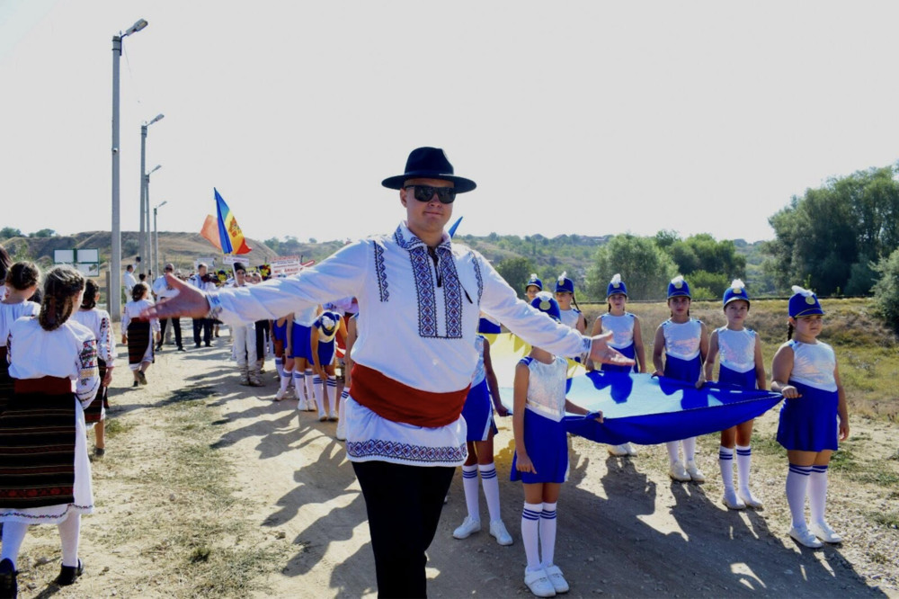 Man in a black hat and white embroidered shirt leads a parade of girls in blue and white outfits on a sunny rural road, holding a blue flag piece.