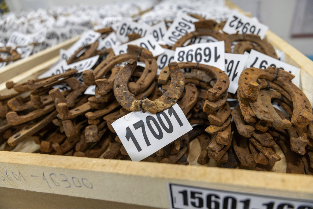 Pile of rusted horseshoes with white numbered tags attached in a wooden crate menagerie of metal, including tag 17001 in focus.