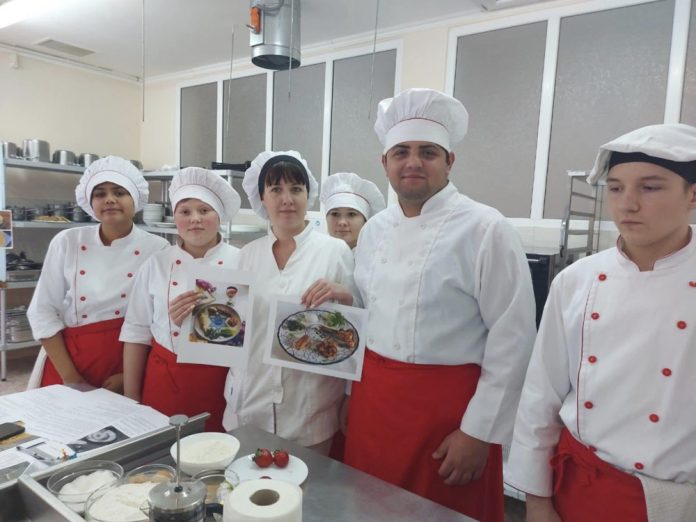 scoala-profesionala Six culinary students in white uniforms and red aprons posing in a kitchen, holding two recipe photos.